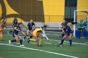 A field hockey match in action, with a player in yellow trying to hit the ball past several players in navy uniforms, as a goalie in blue and green gear stands ready in front of the goal.