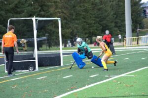 A field hockey player in yellow prepares to shoot at the goal as the goalkeeper in blue and teal blocks the net. An official in orange watches nearby on a green outdoor field.