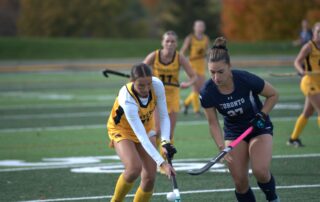 Two women play field hockey on a green field, one in a yellow uniform and the other in navy blue. Other players and autumn trees are visible in the background under a cloudy sky.