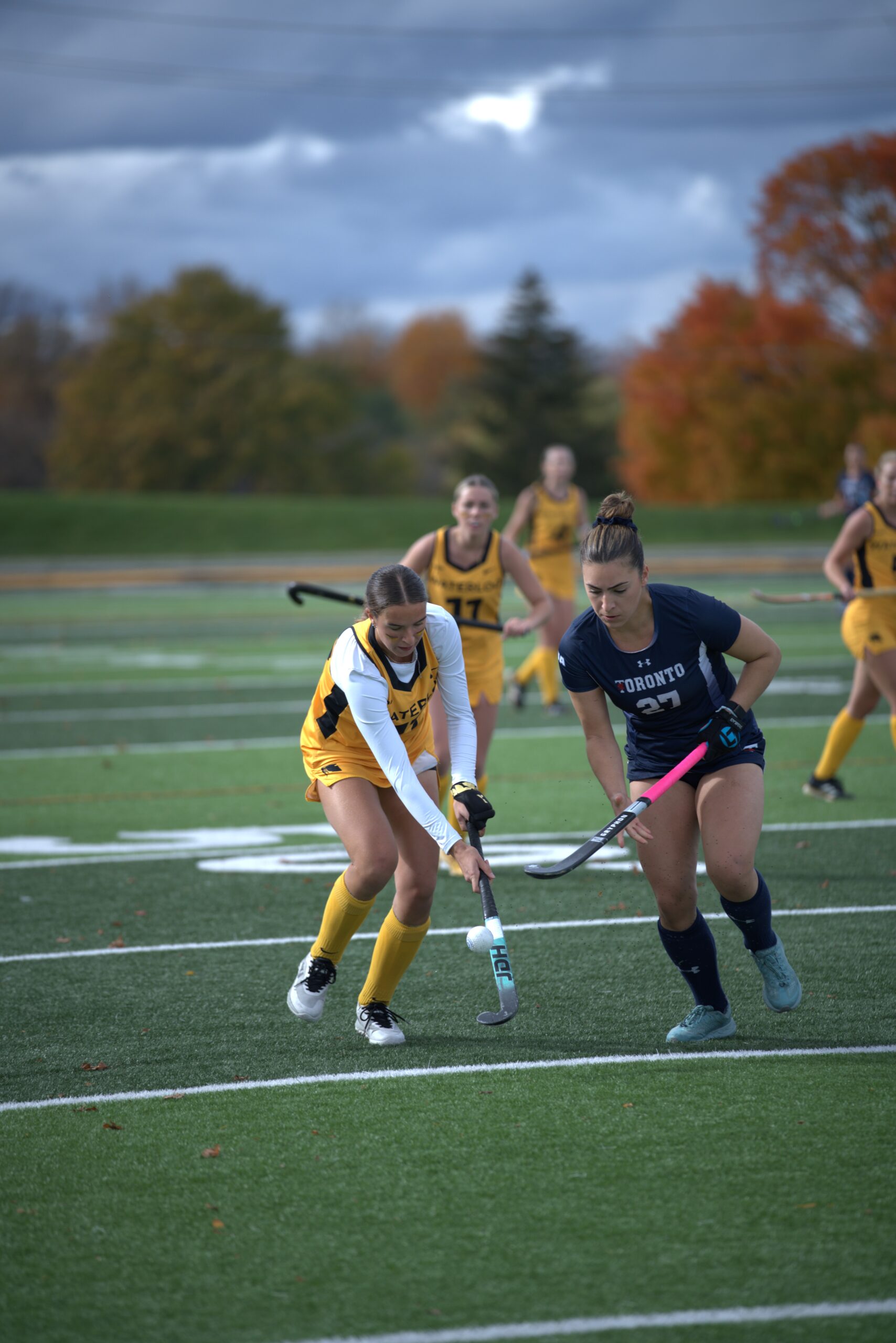 Two women play field hockey on a green field, one in a yellow uniform and the other in navy blue. Other players and autumn trees are visible in the background under a cloudy sky.
