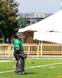 A field hockey goalie, featured among the Athletes of the Week, stands on an outdoor field in protective gear and a green jersey near a white tent and yellow barriers, holding a stick and looking to the side.