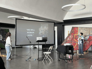 Two people stand in a large room near a projector screen displaying “WE WILL REMAIN HERE,” a powerful message in a space featuring modern art, a podium, and audio-visual equipment—addressing themes of resilience in the face of genocide.