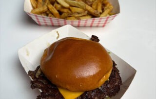 A Bomber cheeseburger with a shiny brioche bun and melted cheese sits in a paper tray, with what I ate—golden French fries—in a separate tray behind it, both on a white table.