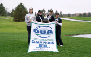 Four women golfers stand on a golf course with their OUA Champions banner and trophy, smiling proudly with OUA gold medals around their necks. All-stars, they celebrate under the trees near a water hazard in the background.