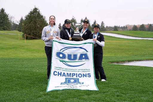 Four women golfers stand on a golf course with their OUA Champions banner and trophy, smiling proudly with OUA gold medals around their necks. All-stars, they celebrate under the trees near a water hazard in the background.