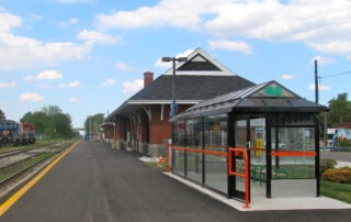 A train station with a brick building, glass shelter with green benches, and train tracks on the left. A Kitchener GO train is parked in the distance under a blue sky with scattered November clouds. Weekend train service is available.