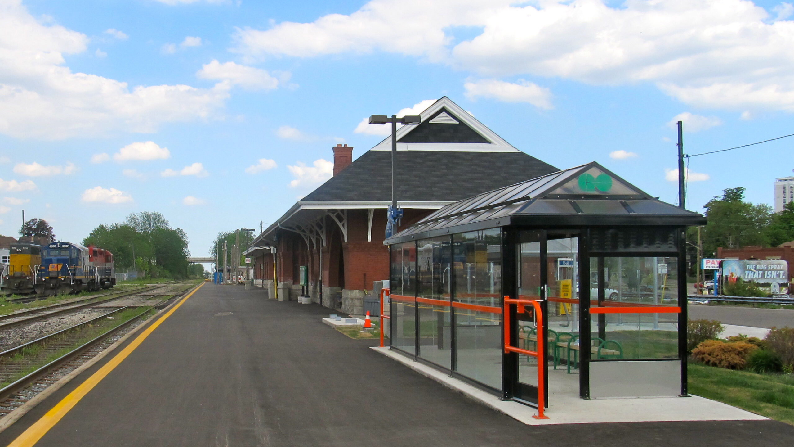 A train station with a brick building, glass shelter with green benches, and train tracks on the left. A Kitchener GO train is parked in the distance under a blue sky with scattered November clouds. Weekend train service is available.