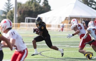 A football player in a black uniform sprints toward the endzone with the ball, chased by players in white and red uniforms on the field. A Warriors team logo is visible in the corner.