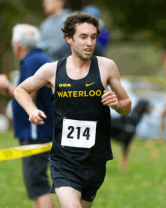 A male runner in a black "WATERLOO" jersey with race number 214 is competing outdoors on a grassy course, highlighted among the athletes of the week, with yellow tape and blurred spectators in the background.