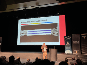 A speaker stands on stage in front of a slide titled "Kaswentha: The Road Forward," showing a blue and white wampum belt—a symbol deeply connected to Indigenous knowledge systems. Banners for the University of Waterloo and Faculty of Mathematics are visible.