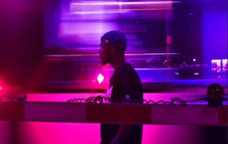 A person wearing a blue cap and dark shirt stands near a red and white barrier, with vibrant pink and purple neon lights swirling as J.Squared music plays in the background, creating a futuristic atmosphere.