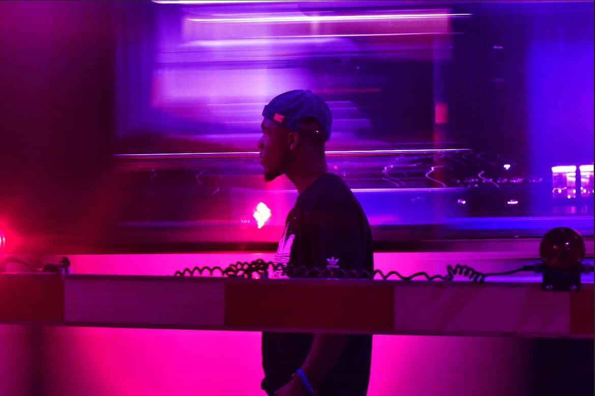 A person wearing a blue cap and dark shirt stands near a red and white barrier, with vibrant pink and purple neon lights swirling as J.Squared music plays in the background, creating a futuristic atmosphere.