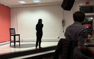 A person in dark clothing and red shoes stands on a small stage with a chair nearby, engaging students in a Politics Without the Podium event, while an audience watches indoors under white walls and overhead lights.