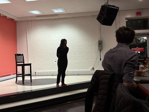 A person in dark clothing and red shoes stands on a small stage with a chair nearby, engaging students in a Politics Without the Podium event, while an audience watches indoors under white walls and overhead lights.