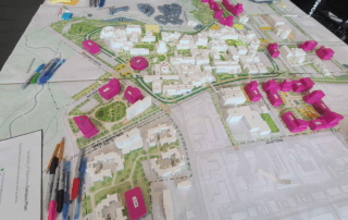A tabletop urban planning map displays a 3D model of a neighborhood, including white buildings and several pink ones. Pens, markers, and papers are arranged around the border for student feedback during the Campus Plan Open House.