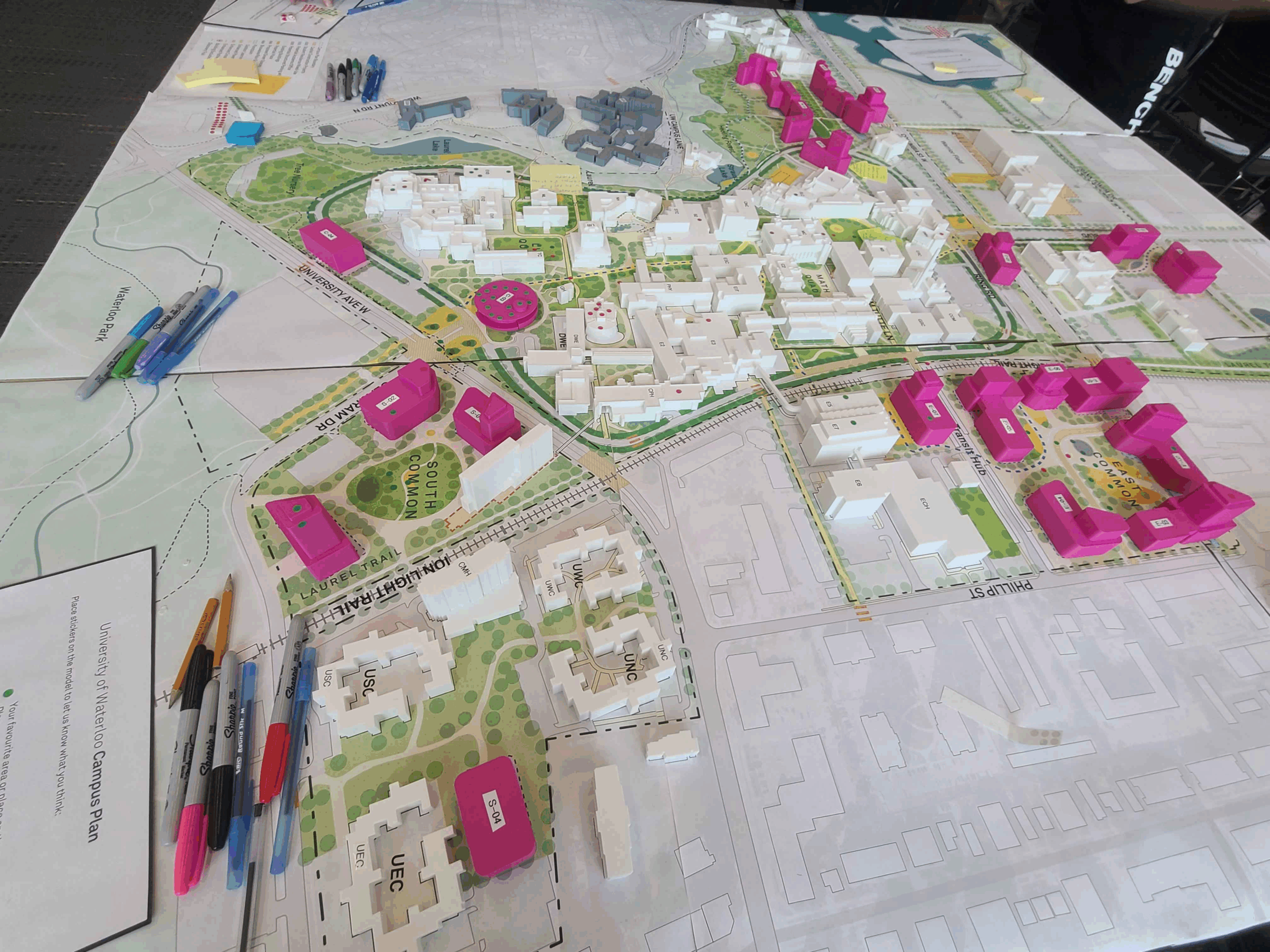A tabletop urban planning map displays a 3D model of a neighborhood, including white buildings and several pink ones. Pens, markers, and papers are arranged around the border for student feedback during the Campus Plan Open House.