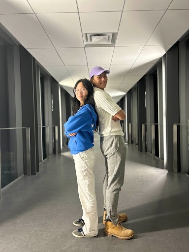 Two people stand back-to-back in a well-lit Waterloo hallway. One wears a blue hoodie and white pants; the other sports a purple cap, white sweater, gray pants, and tan boots. Both have arms crossed and are smiling, ready for their next Wat2Do adventure.