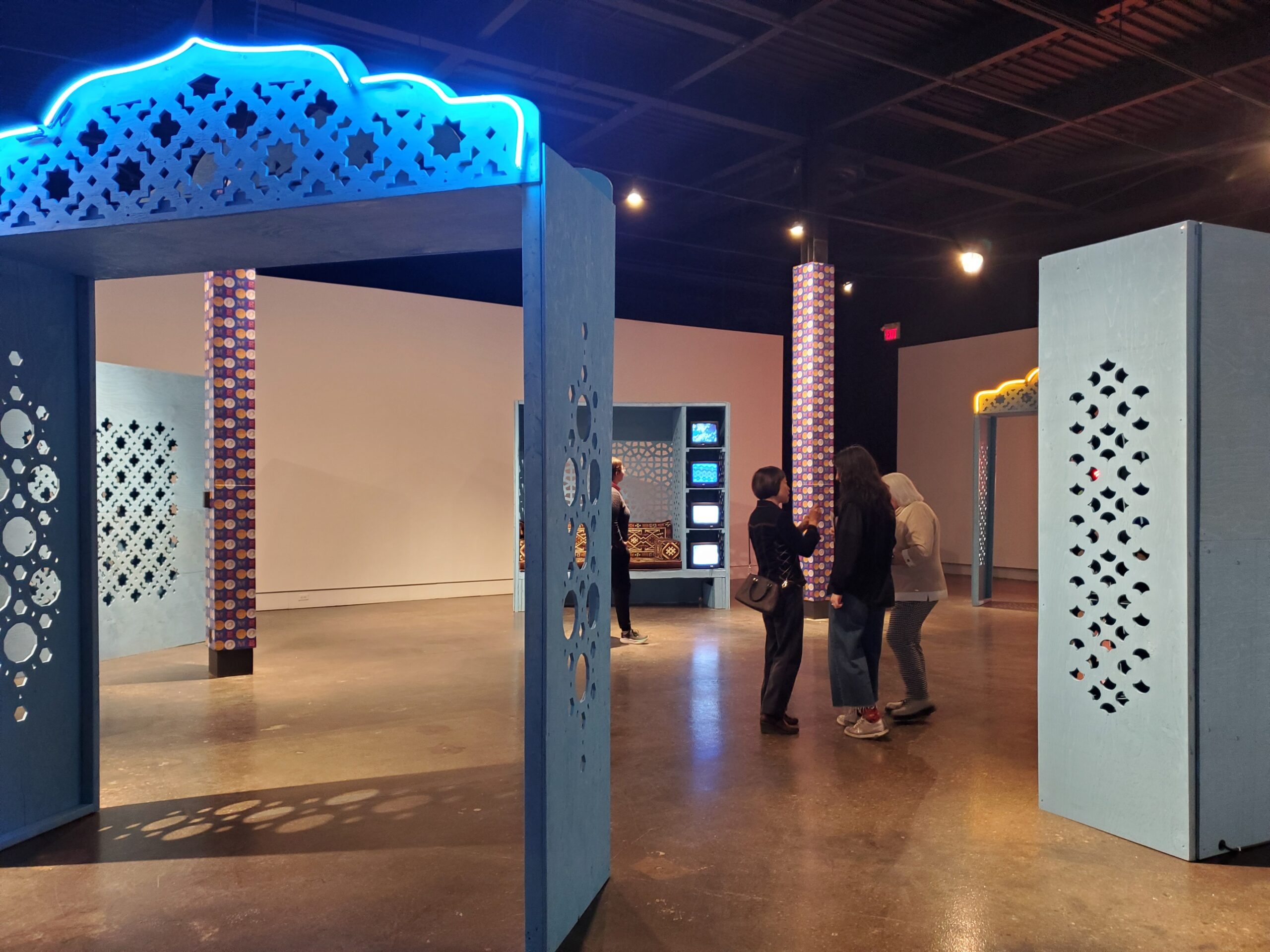 People walk through a UW art exhibition featuring large blue and yellow decorative archways and patterned pillars. Screens display new digital art in the background under warm lighting.