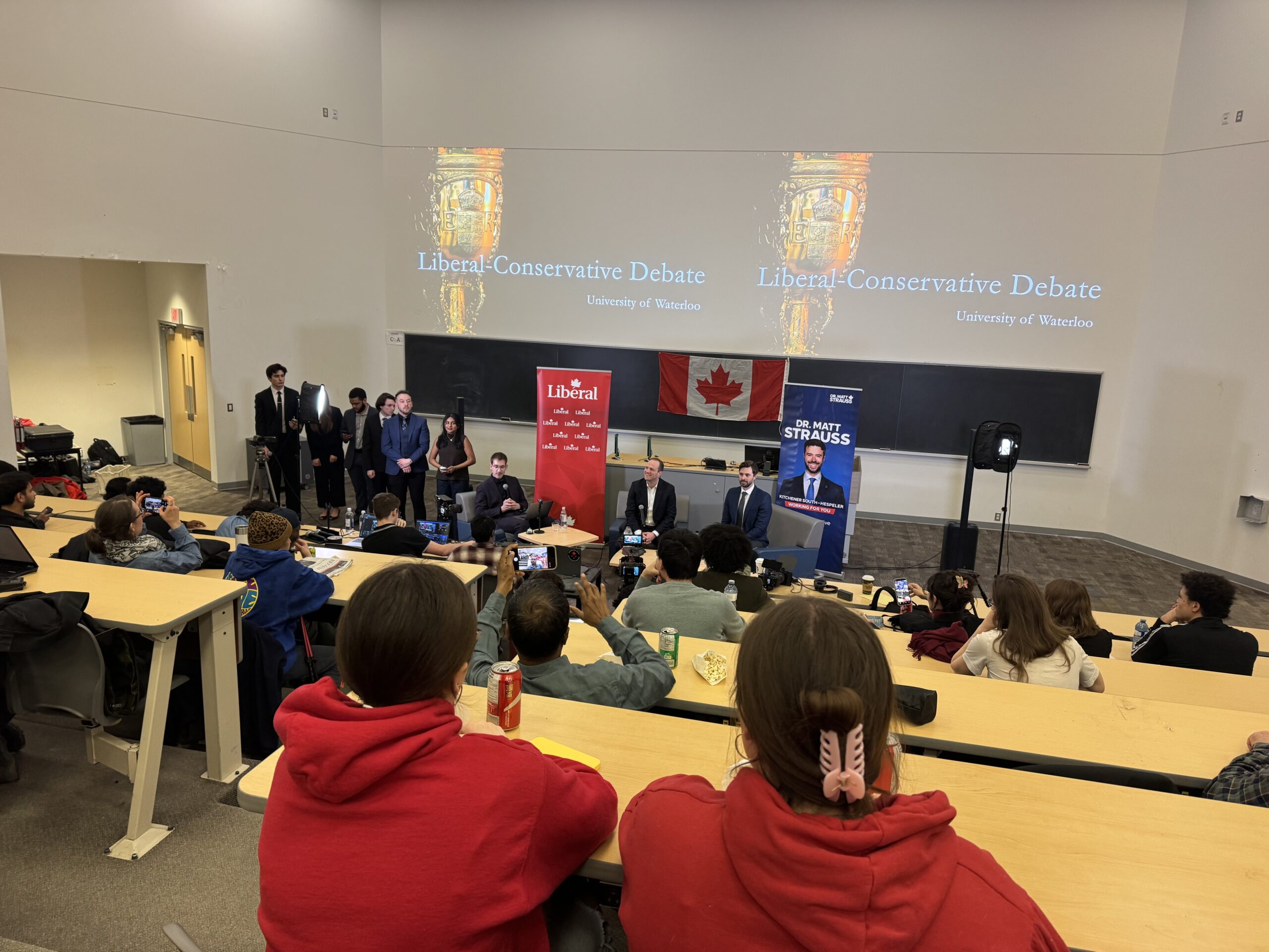 A group of people participate in a Liberal-Conservative debate focused on healthcare and immigration at the University of Waterloo. Audience members sit in a lecture hall, with banners and a Canadian flag displayed at the front.