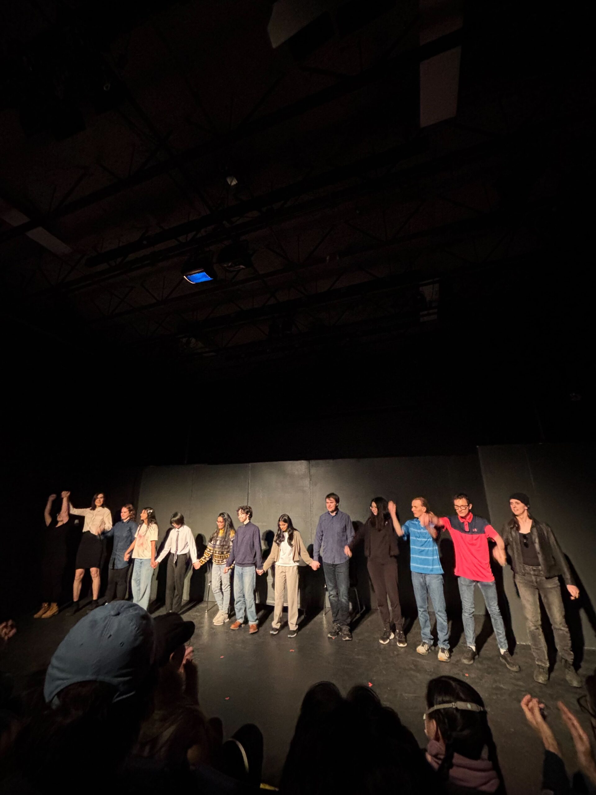 A group of performers, FASS graduates, stands in a line on stage, holding hands and bowing to an applauding audience. The stage is dimly lit with black walls and a high ceiling, capturing a memorable moment after graduation.