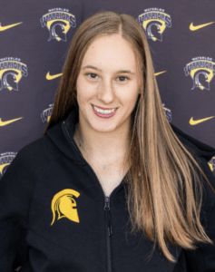 A young woman with straight, light brown hair smiles at the camera. She wears a black jacket with a yellow Spartan helmet logo, celebrating Athletes of the Week. The backdrop features Waterloo Warriors logos and yellow Nike swooshes.