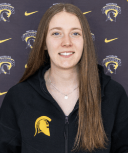 A young woman with straight, long brown hair smiles in front of a backdrop with Waterloo Warriors logos. Keiara Raitt wears a black jacket with a yellow Spartan helmet logo and a silver necklace, celebrating Athletes of the Week honors.