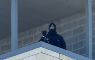A person dressed in black, wearing a mask, stands on a balcony or rooftop of a building—partly hidden by the structure's corner—holding a camera, as if surveying St. Patrick's Day crowds like a WRPS sniper.