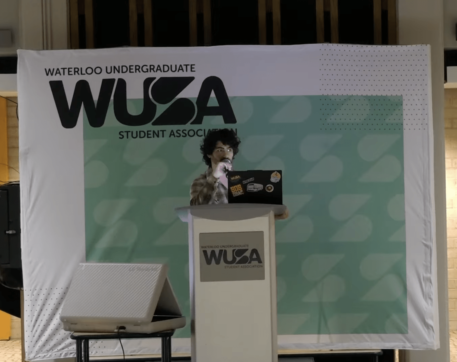 A person stands at a podium with a laptop, speaking into a microphone. The backdrop displays "Waterloo Undergraduate Student Association (WUSA)" in large text, suggesting the speaker may be the WUSA president or one of the vice presidents at an indoor event.