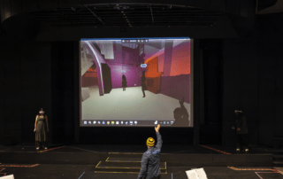 During a UW Theatre performance, a person on stage points at a large screen showing a virtual scene from The Nether, featuring human figures in a brightly colored, geometric environment as two others stand by mannequins or props.