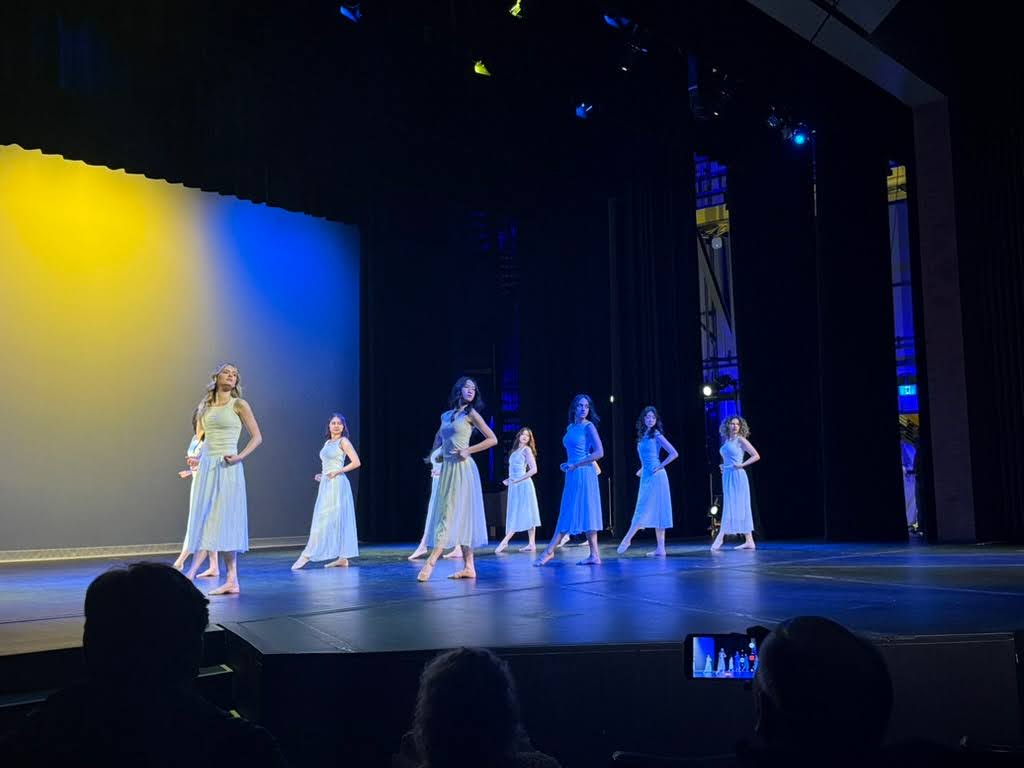 Seven dancers in flowing white and light blue dresses perform on stage, their movements telling tales through wearable art. Dramatic lighting bathes the scene in blue and yellow, while audience silhouettes watch this Fashion for Change moment unfold.
