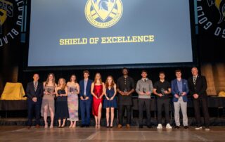 A group of twelve people dressed formally stand on stage, each holding an award. Behind them, a large screen displays a Warrior logo and the text "Shield of Excellence.