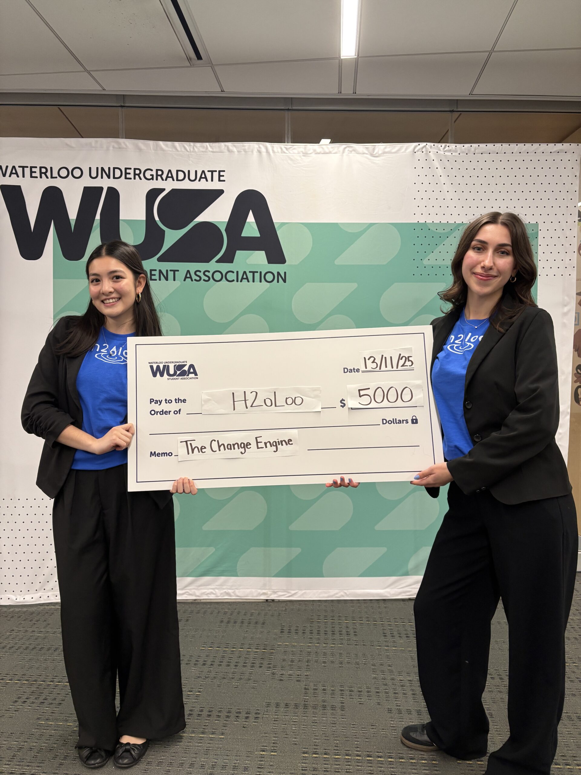 Two UW students smile while holding a large cheque for $5,000 from the Waterloo Undergraduate Student Association, awarded to H2oLoo for "The Change Engine." The background features a patterned wall and WUSA logo.