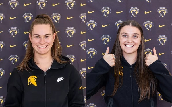 Two young women, Nikki McDonald and Tatum James, in black jackets with yellow sports logos, smile before a backdrop of gold warrior helmets and Nike swooshes. One gives two thumbs up—both are proud Athletes of the Week.