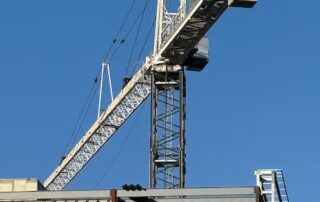 A tall construction crane extends above a partly built concrete and steel building against a clear blue sky. Shadows from the crane and structure highlight areas designated for temporary closures during crane removal.