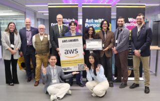 A group of twelve UW students pose indoors, some standing and some kneeling, holding an AWS Awards sign, a large check for their municipal service app, and a QR code. Banners behind them read, “Think Different,” “Act with Purpose,” and “Work Together.”.