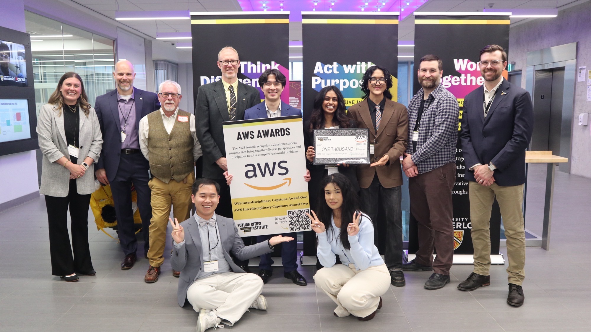 A group of twelve UW students pose indoors, some standing and some kneeling, holding an AWS Awards sign, a large check for their municipal service app, and a QR code. Banners behind them read, “Think Different,” “Act with Purpose,” and “Work Together.”.