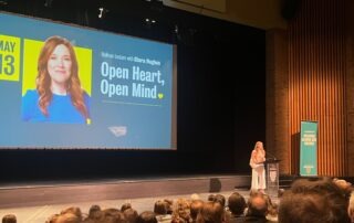 A woman stands at a podium in a UW auditorium, speaking to an audience. Behind her, a screen displays "Hahman Lecture with Clara Hughes: Open Heart, Open Mind," highlighting her insights on mental health.