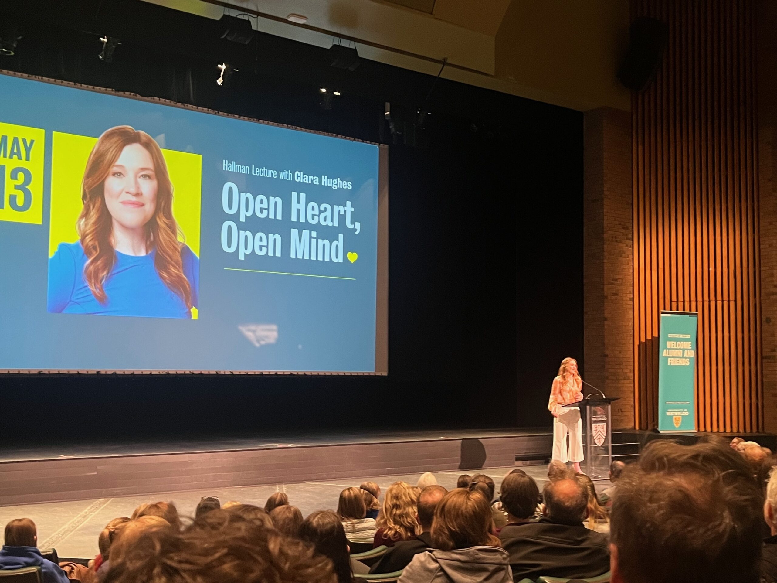 A woman stands at a podium in a UW auditorium, speaking to an audience. Behind her, a screen displays "Hahman Lecture with Clara Hughes: Open Heart, Open Mind," highlighting her insights on mental health.