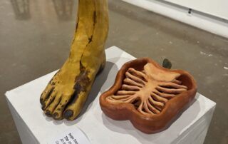 Two ceramic sculptures on a white pedestal: a yellow banana shaped like a human foot, and a sliced bell pepper with the inside carved to look like a human rib cage.