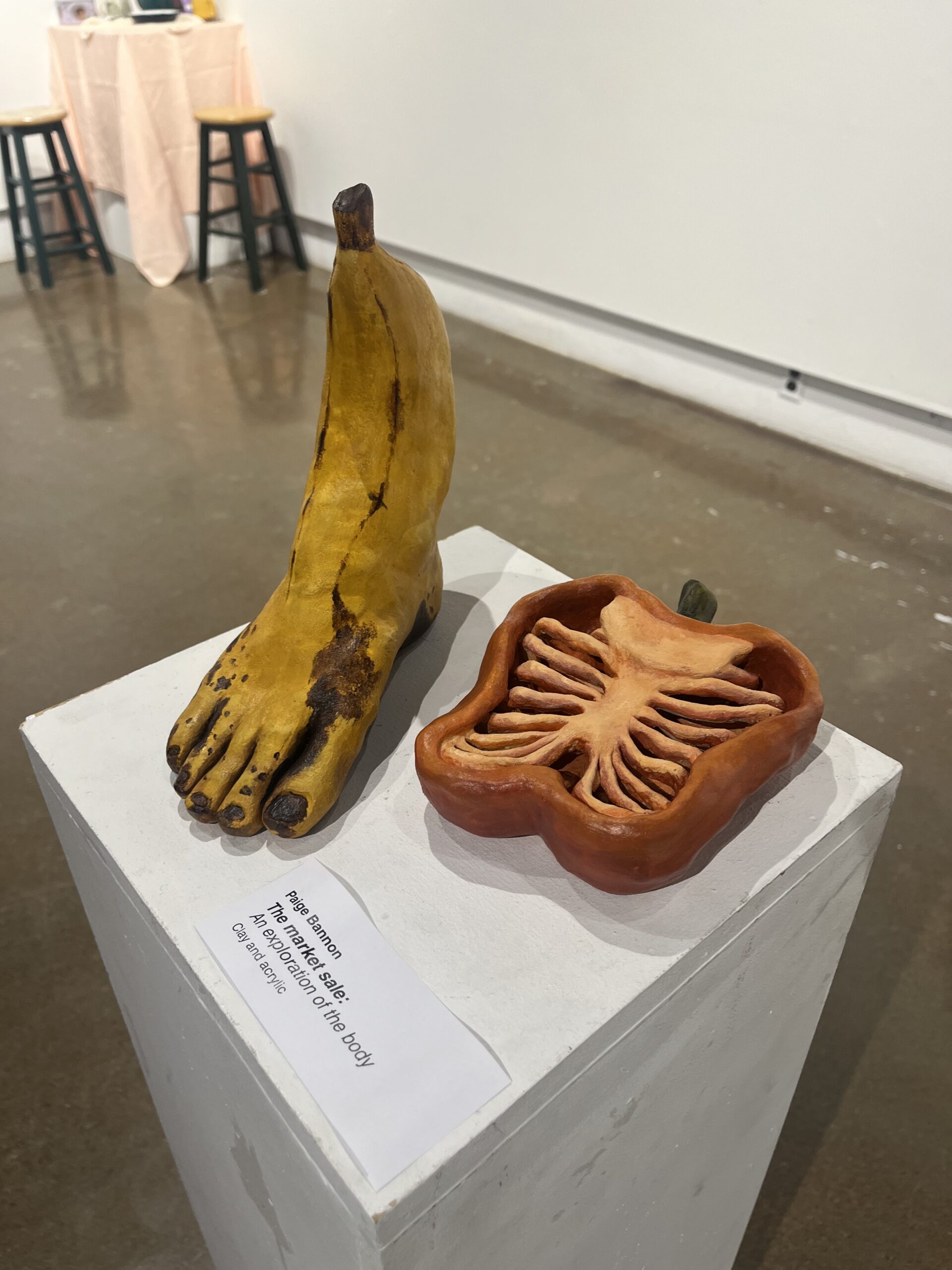 Two ceramic sculptures on a white pedestal: a yellow banana shaped like a human foot, and a sliced bell pepper with the inside carved to look like a human rib cage.