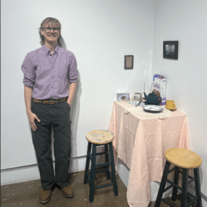A blonde man wearing glasses, a purple shirt, and dark pants stands smiling next to a small table covered with a pink cloth, two stools, dishes, a mug, and a box of Cheerios at the SOFA starving art exhibit. Picture frames hang on the white wall behind the table.