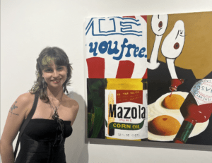 A person with short, wavy hair and a black dress stands smiling beside a painting at the SOFA art exhibit, featuring Mazola corn oil, wine, and the words "you free".