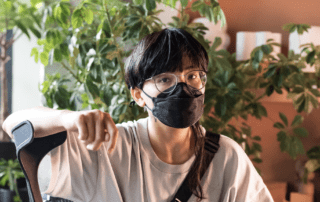 A masked non-binary Filipino person looks at the camera while sitting with his legs crossed, one arm casually leaning on a forearm crutch. He wears glasses, a face mask, a loose fit T-shirt, long shorts, and hair that’s short and blunt except for a single long strand. Indoor plants and pots fill the background.
