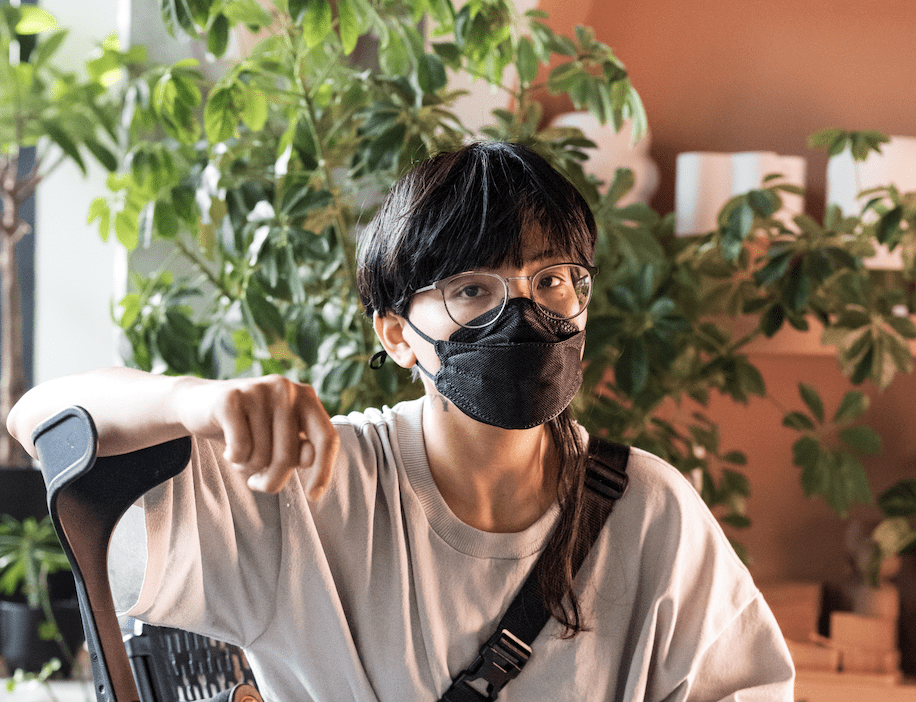 A masked non-binary Filipino person looks at the camera while sitting with his legs crossed, one arm casually leaning on a forearm crutch. He wears glasses, a face mask, a loose fit T-shirt, long shorts, and hair that’s short and blunt except for a single long strand. Indoor plants and pots fill the background.