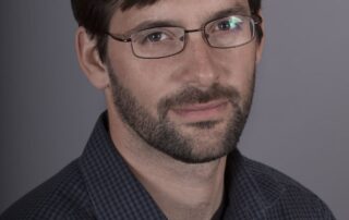 A man with short brown hair, a beard, and glasses is wearing a dark, checkered shirt and looking at the camera with a neutral expression. The background is plain and gray. He resembles psychology professor Daniel Smilek, recipient of the Distinguished Teacher's Award.