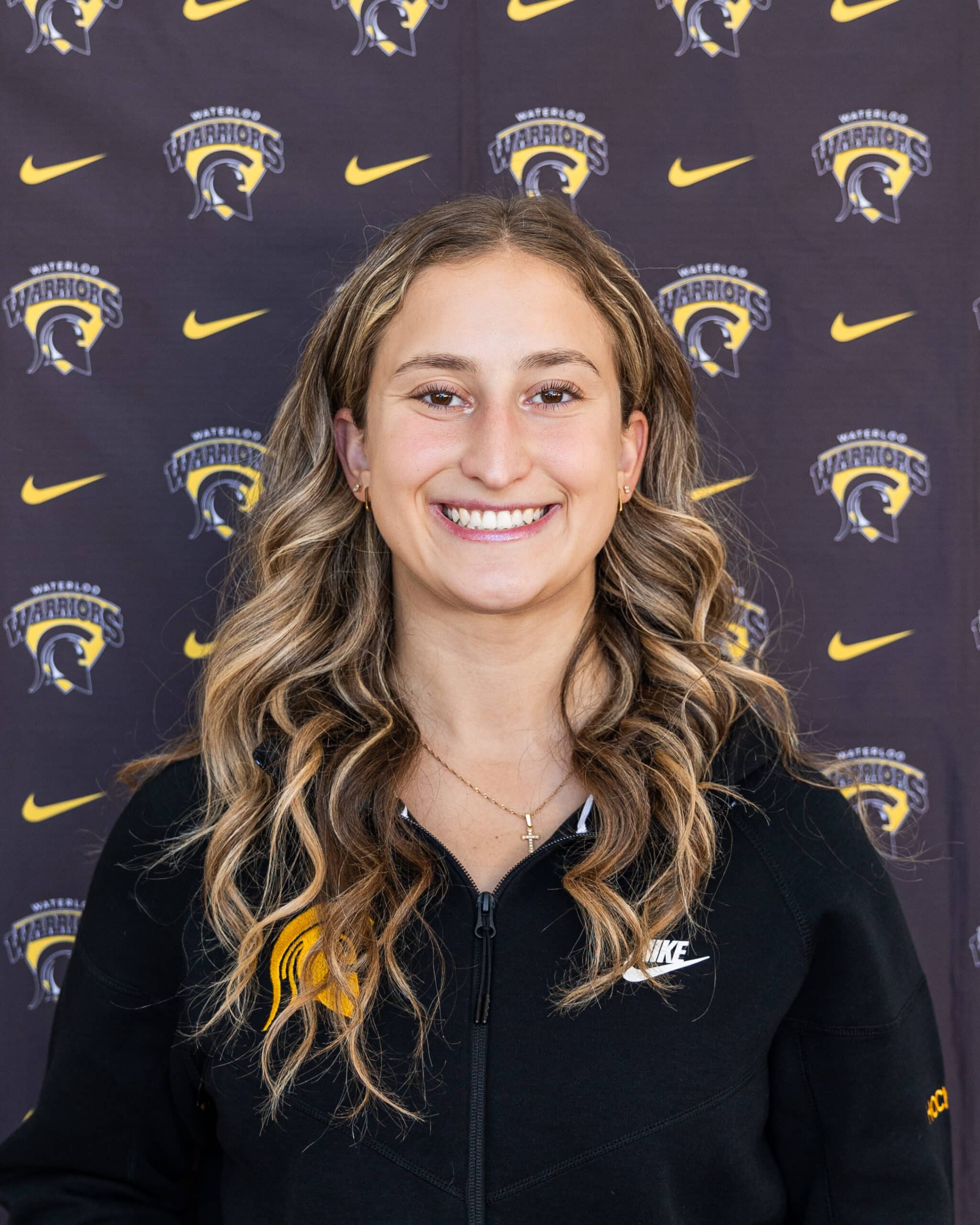 Smiling young woman with wavy hair stands in front of a backdrop featuring Waterloo Warriors and Nike logos. A proud Warriors hockey player, she is wearing a black jacket with the Warriors logo, celebrating her hockey career.
