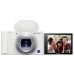 A white Sony digital camera from Best Buy, featuring a furry microphone and a flip-out screen, captures three smiling friends taking a selfie outdoors—an ideal Back to School Tech pick for memorable moments.