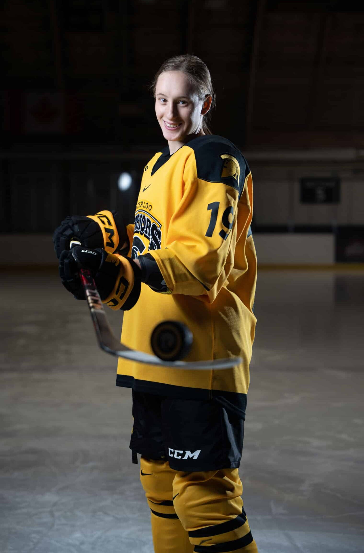 Katina Duscio in a yellow and black jersey stands on an ice rink holding a hockey stick with a puck balanced on the blade.