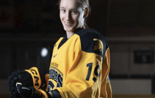 Katina Duscio in a yellow and black jersey stands on an ice rink holding a hockey stick with a puck balanced on the blade.