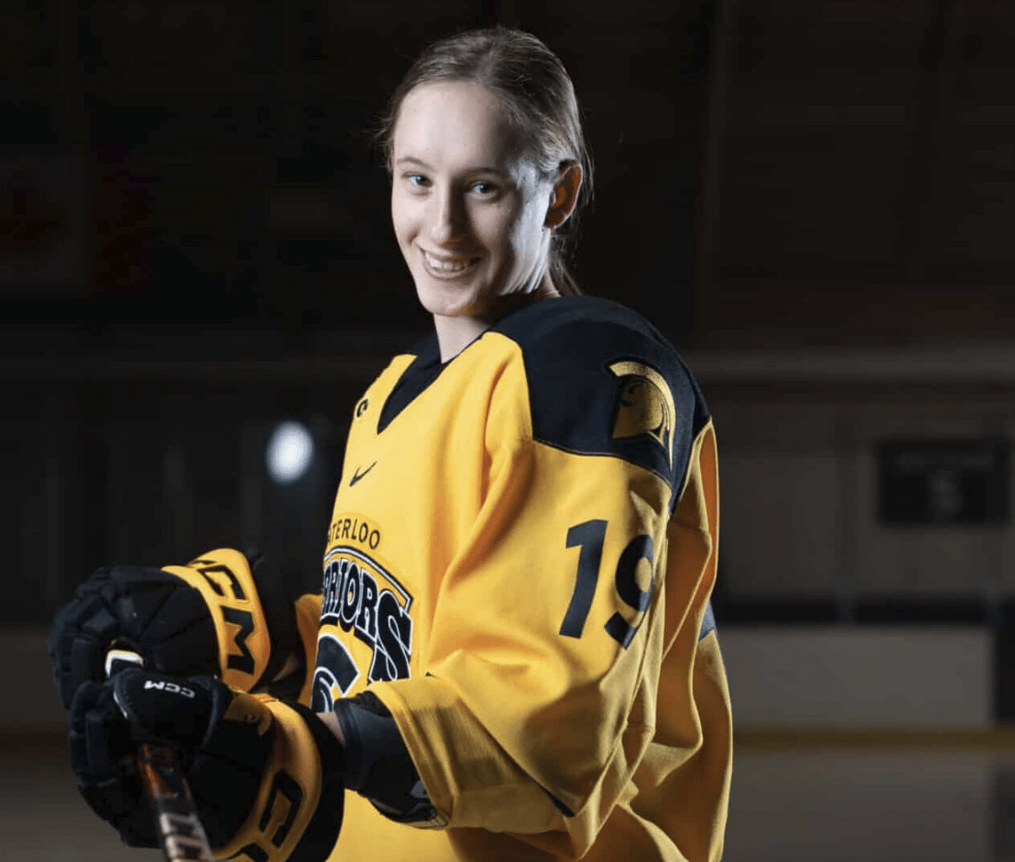 Katina Duscio in a yellow and black jersey stands on an ice rink holding a hockey stick with a puck balanced on the blade.